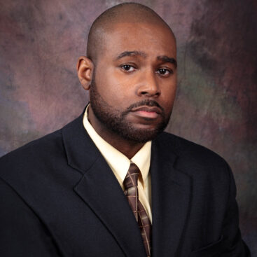 Professional portrait of a man in a dark suit and tie against a textured background.
