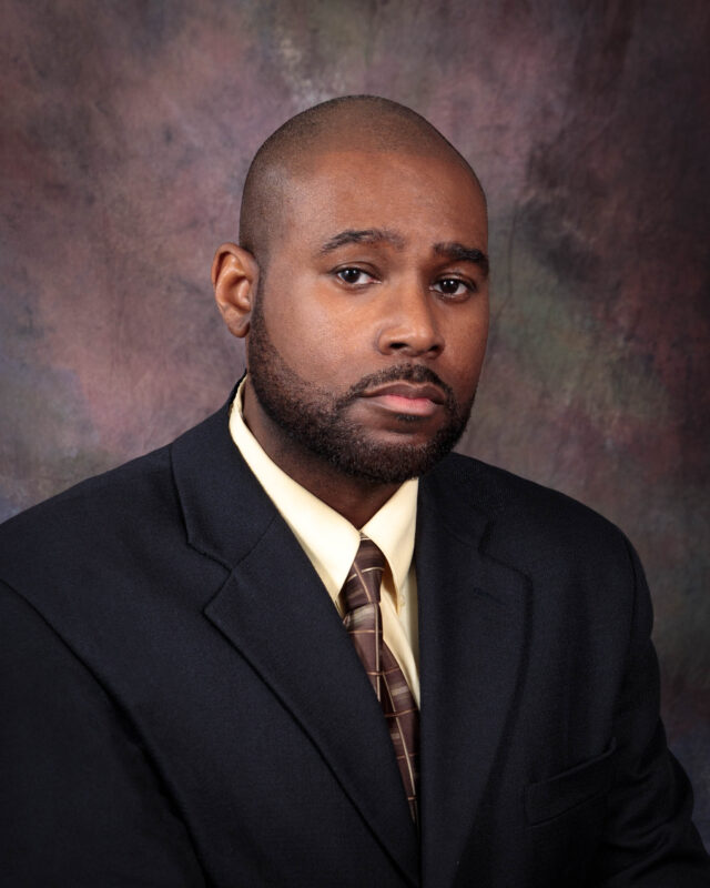 Professional portrait of a man in a dark suit and tie against a textured background.