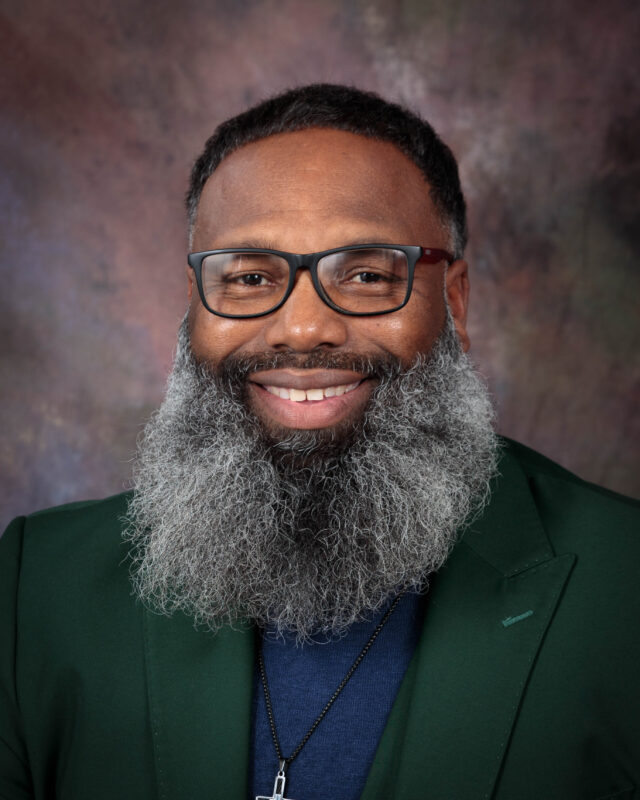 Smiling man with a full gray beard and glasses in a formal suit.